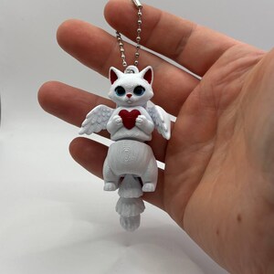 Könnte beinhalten: Eine weiße Katzenfigur mit Engelsflügeln und einem roten Herzen. Die Katze hat blaue Augen und eine Kette zum Aufhängen. Die Figur besteht aus einem 3D-gedruckten Material.