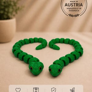 Op de afbeelding: Twee gearticuleerde groene slangen met geel-zwarte ogen, 3D-geprint met een gesegmenteerd lichaam. De afbeelding bevat een cirkelvormig embleem met de tekst MADE IN AUSTRIA en een onderste paneel met iconen en de tekst: MIT LIEBE GEMACHT, 3D GEDRUCKT IN ÖSTERREICH, HOCHWERTIGE QUALITÄT, NACHHALTIG & UMWELTFREUNDLICH.