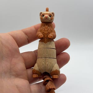 Könnte beinhalten: Segmentierte Frettchenfigur in Braun- und Beigetönen. Kopf und Schwanz sind dunkelbraun, während der Körper hellbeige ist. Die Figur wird in einer Hand vor weißem Hintergrund gehalten.