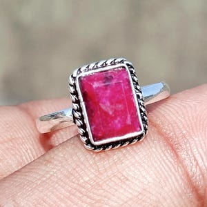 May include: A silver ring featuring a rectangular, vibrant pink gemstone set in a twisted rope-style metal border. The band is smooth and polished, contrasting with the textured setting that frames the faceted, deep magenta stone at the center.
