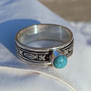 May include: A silver ring with a turquoise stone. The ring has a detailed band with floral and geometric patterns. The turquoise stone is round and set in a silver bezel. The ring is displayed on a white surface.