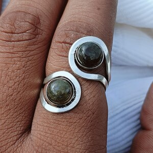 May include: A bypass ring featuring two round, dark green cabochon stones set in a polished silver-toned metal band. The design consists of two open, swirling ends that each hold a single circular gemstone, creating a continuous, open-ended look.
