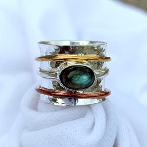 May include: A wide silver spinner ring featuring a hammered texture, a central oval labradorite gemstone with blue-green iridescence, and two thin rotating bands in gold and copper tones that wrap around the main silver band.