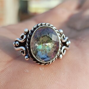 May include: Silver-coloured ring featuring an oval, iridescent gemstone. The stone reflects green, blue, and purple hues. The setting is ornate, with decorative swirls and a rope-style border. The ring is a piece of jewellery.