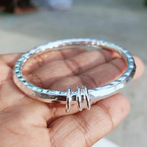May include: A silver-colored bangle bracelet with a hammered, textured surface. The circular band features three small, smooth silver-colored rings that are looped around the main body of the bracelet, creating a distinct focal point.