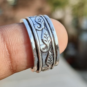 May include: A silver-colored ring with a detailed floral and scroll design. The ring has a spinning band and is held on a finger. The ring's design includes leaves and swirling patterns, creating a textured appearance.