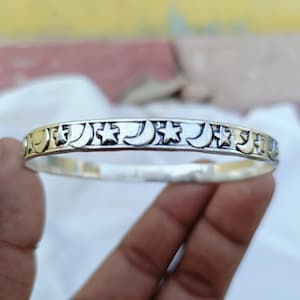 Könnte beinhalten: Ein silberfarbenes Armreif-Armband mit einem sich wiederholenden Muster aus Monden und Sternen. Das Armband ist kreisförmig und hat eine polierte Oberfläche. Das Design ist in das Metall geätzt und erzeugt ein detailliertes Himmelsmotiv.