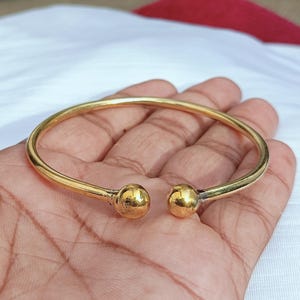 May include: A gold-toned metal cuff bracelet featuring a smooth, circular band that remains open at the front. The two ends of the bracelet are finished with polished, spherical ball terminals, creating a simple and continuous metallic design.