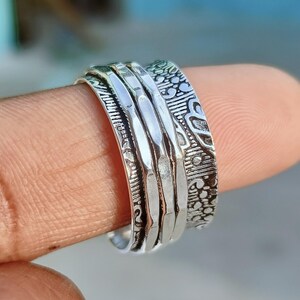 May include: A silver-colored spinner ring with intricate etched designs. The ring features multiple bands, including textured and patterned sections. The ring is shown on a finger, highlighting the details of the jewelry.