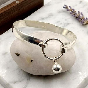 May include: A silver bangle bracelet featuring a circular clasp and a small, dangling sphere. The bracelet is presented on a light-coloured stone against a marble surface, with a wooden box and lavender sprig in the background.