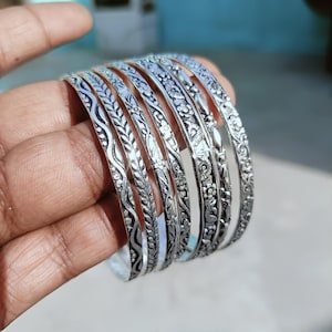 May include: A collection of silver bangles with intricate floral and leaf patterns. The bangles are displayed in a hand, showcasing the detailed designs and the reflective quality of the metal. The bangles are a beautiful accessory.