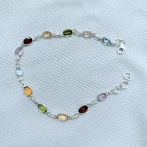 May include: A silver bracelet featuring a series of oval-shaped gemstones in various colors, including red, green, yellow, and blue. The gemstones are linked together by a delicate silver chain, creating a colorful and elegant accessory.