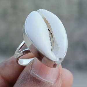 May include: A silver ring featuring a large, white cowrie shell. The shell has a natural, textured appearance with a central opening. The ring's band is a simple, split design, and the setting is polished silver.