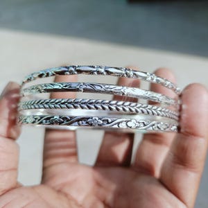May include: A set of five silver bangles with intricate floral and geometric patterns. The bangles are held in a hand, showcasing the detailed designs and the reflective quality of the metal. Each bangle has a unique pattern.
