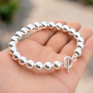 May include: A silver bracelet featuring large, round beads. The bracelet has a toggle clasp and is displayed on a person's hand. The beads are highly polished, reflecting light, and the overall design is simple and elegant.