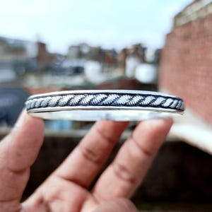 Op de afbeelding: Een zilverkleurige armband met een gevlochten touwontwerp in het midden. De armband wordt in een hand gehouden, met een vage achtergrond van gebouwen en lucht.