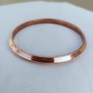 May include: A solid copper bangle bracelet with a smooth, polished finish and a slightly angled, beveled edge. The circular metal band features a warm, metallic reddish-brown hue with subtle light reflections across its continuous, seamless surface.
