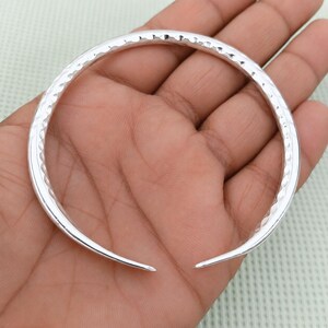 Op de afbeelding: Een zilverkleurige, open armband met een gehamerde textuur. De armband wordt in een hand gehouden, waarbij de gepolijste afwerking en het eenvoudige ontwerp worden benadrukt. Een elegant sieraad voor dagelijks gebruik.