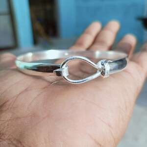 May include: A silver-colored bangle bracelet with a hook-and-eye clasp. The bracelet is round and smooth, with a polished finish. The clasp is a decorative element, adding visual interest to the jewelry.