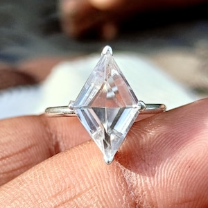 May include: A silver ring featuring a large, clear, kite-shaped gemstone set in a four-prong setting. The stone has a faceted surface that reflects light, and the band is a simple, smooth, polished silver metal that encircles the finger.