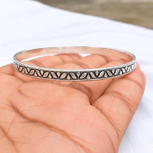 May include: A silver-toned bangle bracelet featuring a continuous embossed pattern of small, stylized leaves around the entire outer circumference. The metal has a polished finish with darkened recessed areas that highlight the leaf design details.