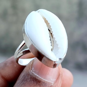 May include: A ring featuring a polished white cowrie shell set in a smooth silver-toned metal bezel. The shell has a characteristic central slit with serrated edges. The band is made of a simple, thin silver-toned metal wire that splits into two.