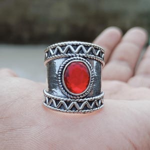 Op de afbeelding: Een zilverkleurige ring met een grote, ovale, rode edelsteen in het midden. De ring heeft een brede band met decoratieve zigzag- en touwachtige patronen. De edelsteen is gezet in een touwachtige rand.
