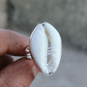 May include: A silver ring featuring a large, white cowrie shell as its centerpiece. The shell is oval-shaped with a natural, textured surface. The ring band is made of multiple silver bands.