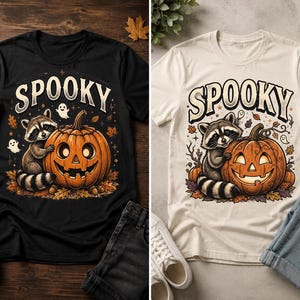 May include: A black and a cream-coloured t-shirt, each featuring a graphic print of a raccoon hugging a jack-o'-lantern pumpkin surrounded by autumn leaves and small ghosts, with the word SPOOKY printed in large, stylized lettering above the design.