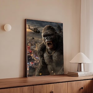 May include: A framed print depicts a large gorilla roaring above a city skyline, with helicopters in the sky. The artwork features a gold chain around the gorilla's neck and is set against a backdrop of a sunset.