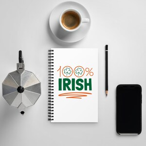 Könnte beinhalten: Ein weißes Notizbuch mit dem Text "100% Irish" in Grün und Orange, mit Kleeblattsymbolen. Eine Tasse Kaffee, ein Bleistift, ein Smartphone und eine silberne Kaffeemaschine befinden sich ebenfalls auf der weißen Oberfläche.