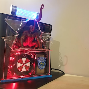 May include: Custom computer case with a Resident Evil theme. It has a red and black colour scheme, a glowing blue tube, and a monster figure. Includes the Umbrella Corporation logo and S.T.A.R.S. badge.