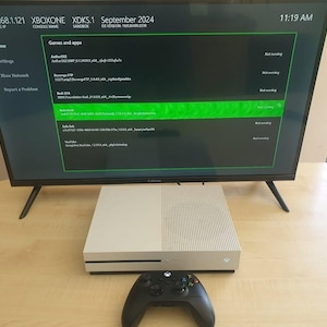 Puede incluir: Un televisor de pantalla plana negro que muestra una interfaz verde y negra, con el texto "XBOXONE" y "Septiembre 2024". Debajo, una consola de juegos blanca y un mando negro.