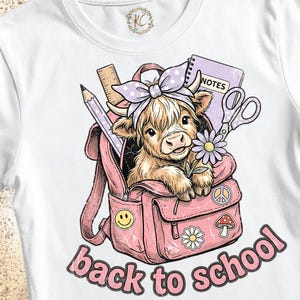 May include: A white t-shirt featuring a graphic of a fluffy highland cow wearing a purple polka dot bow, sitting inside a pink backpack. The backpack contains a ruler, pencil, and notebook labeled NOTES. Below, the text reads back to school.