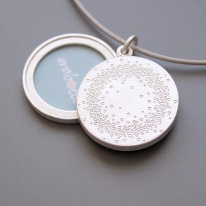 Puede incluir: Un collar con un medallón de plata con un diseño circular en la parte delantera y un pequeño marco de fotos en la parte posterior. El medallón está abierto, revelando un espacio en blanco para una foto.