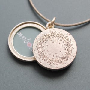 Könnte beinhalten: Silberne Halskette mit Anhänger, auf der Rückseite mit einem kreisförmigen Design. Die Vorderseite des Anhängers ist offen und zeigt ein kleines Stück Papier mit dem Wort "mabo" darauf.