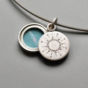 May include: Silver locket necklace with a sunburst design on the back. The locket opens to reveal a small space for a photo or message. The necklace is on a gray cord.
