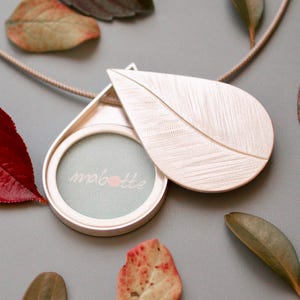 Puede incluir: Un collar de plata con un dije en forma de hoja, con una abertura redonda que revela un fondo azul claro con la palabra "mabottle" escrita en blanco. El dije está abierto y la parte posterior del dije es visible.