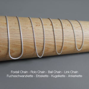May include: A collection of silver necklaces displayed on a wooden dowel. The necklaces include foxtail, rolo, ball, and link chain styles. The image also includes the text "Foxtail Chain - Rolo Chain - Ball Chain - Link Chain".