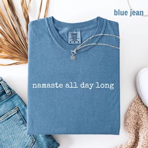 May include: A blue jean colored t-shirt with the text namaste all day long printed in white across the chest. The shirt features a crew neckline and a Comfort Colors brand label on the inside collar. A silver chain necklace rests on the shirt.