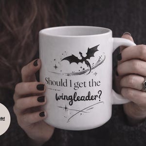 Puede incluir: Taza de cerámica blanca con una ilustración de dragón negro y el texto "Should I get the wingleader?" La taza es sostenida por una persona. La taza es un regalo perfecto.