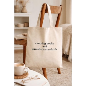 May include: A beige tote bag with the text "carrying books and unrealistic standards" in black. The bag is hanging on a wooden chair. A cup of coffee, glasses, and a notebook are on a table in front of the chair.