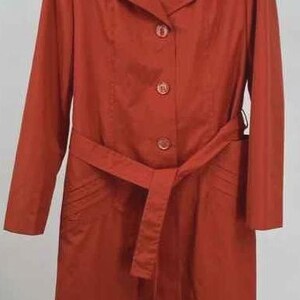 May include: A long, rust-colored trench coat with a matching belt. The coat features a classic collar, button-down front, and long sleeves. The coat is hanging on a wooden hanger.