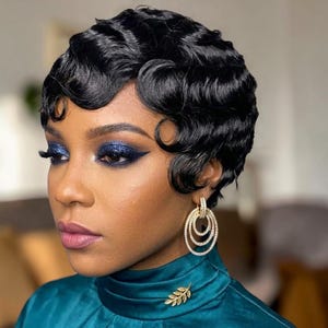 Könnte beinhalten: Eine Frau mit dunkler Haut und einer kurzen, welligen schwarzen Frisur. Sie trägt blauen Lidschatten, Eyeliner und rosa Lippenstift. Goldene Ohrringe und eine goldene Blattbrosche schmücken sie. Sie trägt ein türkisfarbenes Oberteil.
