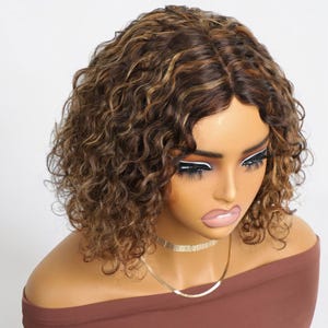 May include: A brown curly wig with blonde highlights. The wig is styled in a short, bob-like cut. The wig is displayed on a mannequin head with a brown top and a gold necklace.