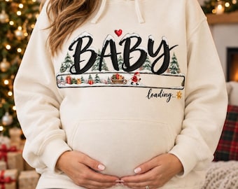 Peut inclure: Un sweat-shirt à capuche couleur crème avec une impression graphique comportant le texte BABY en grandes lettres noires, un petit cœur rouge et une barre de progression illustrée avec des sapins, des cadeaux, un traîneau et le Père Noël, avec le texte loading... et une étoile dorée.
