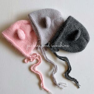 May include: Three knitted baby hats with ear flaps and ties. The hats are pink, gray, and dark gray.