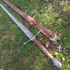 Könnte beinhalten: Ein Langschwert mit gemusterter Stahlklinge, einem Parierstange mit kleinem grünen Stein und einem braunen, lederumwickelten Griff. Dazu gehört eine passende braune Lederscheide mit geprägten Verzierungen und einer Gürtelhalterung.