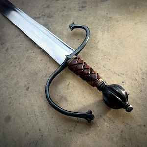 Könnte beinhalten: Ein Schwert mit einer langen, polierten Stahlklinge und einem dunklen, geschwungenen Metall-Parierstange. Der Griff besteht aus einem strukturierten, rotbraunen Material mit Rautenmuster und endet in einem runden, dunklen Metallknauf.