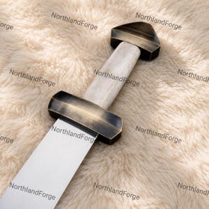 Könnte beinhalten: Ein Schwert mit einer silberfarbenen Metallklinge, einem Parierstange und Knauf in dunkler, gebürsteter Bronzeoptik sowie einem hellen, strukturierten Griff. Der Text NorthlandForge ist in dunkler Schrift über das Bild verteilt.